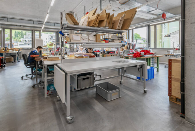 Table d'emballage ERGO-Basic équipée de roulettes, dans un atelier lumineux doté de rayonnages de stockage et où travaillent des employés